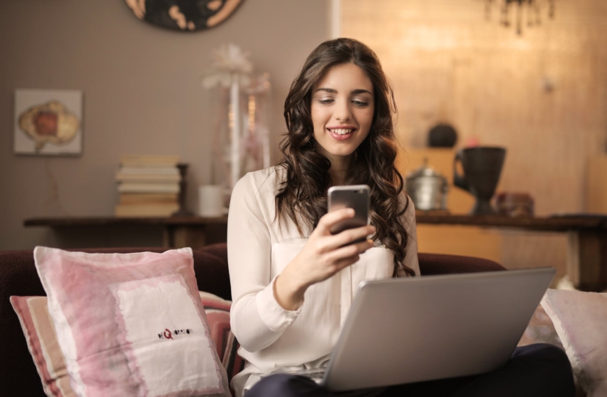 woman sitting on sofa while looking at phone with laptop on lap
