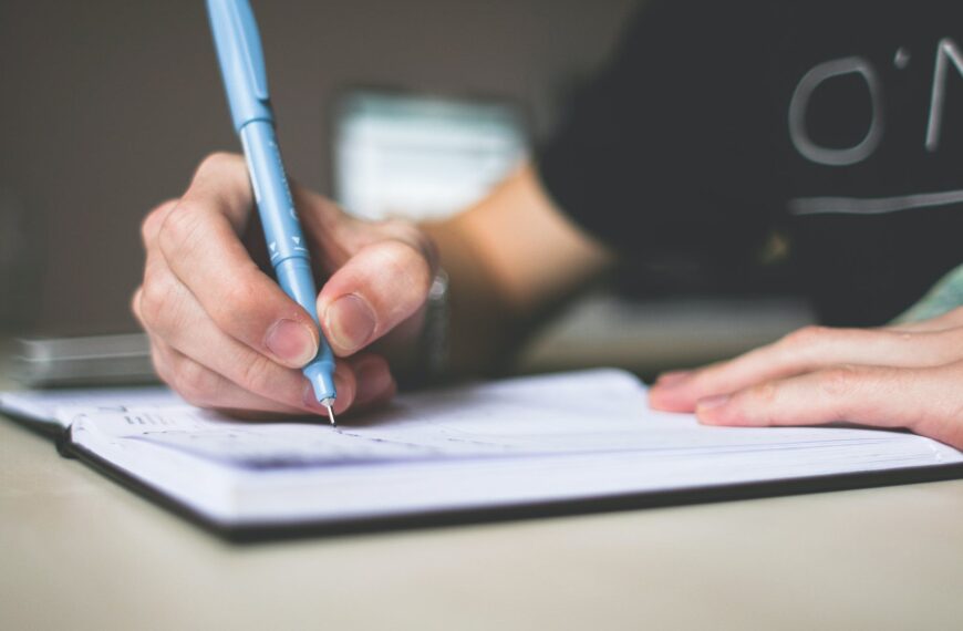 person holding blue ballpoint pen writing in notebook