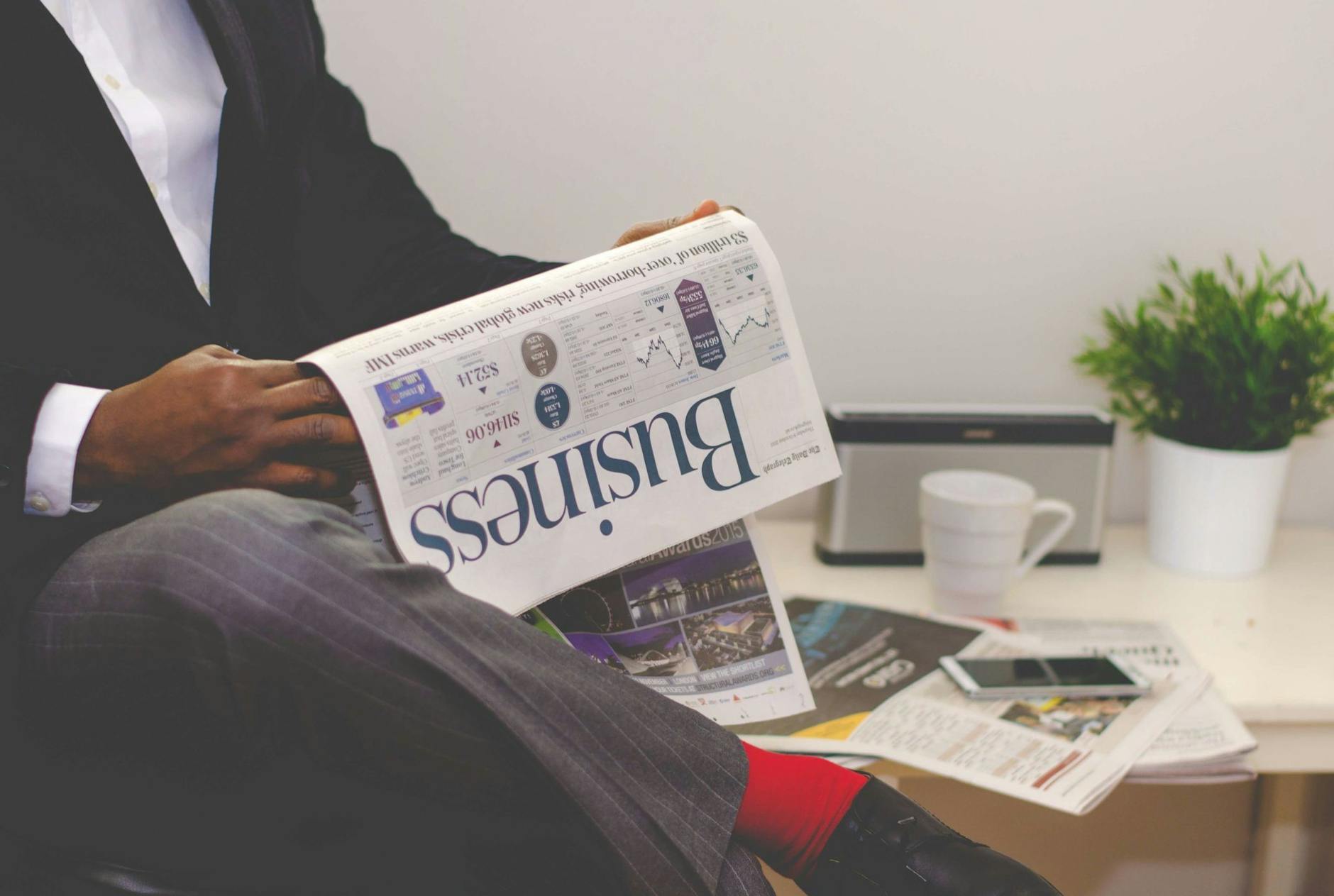 man reading newspaper while sitting near table with smartphone and cup